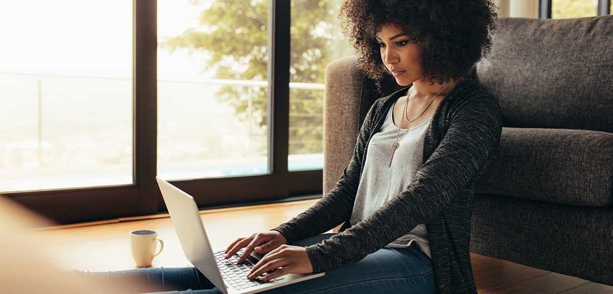 Online student sits in front of couch, works on laptop