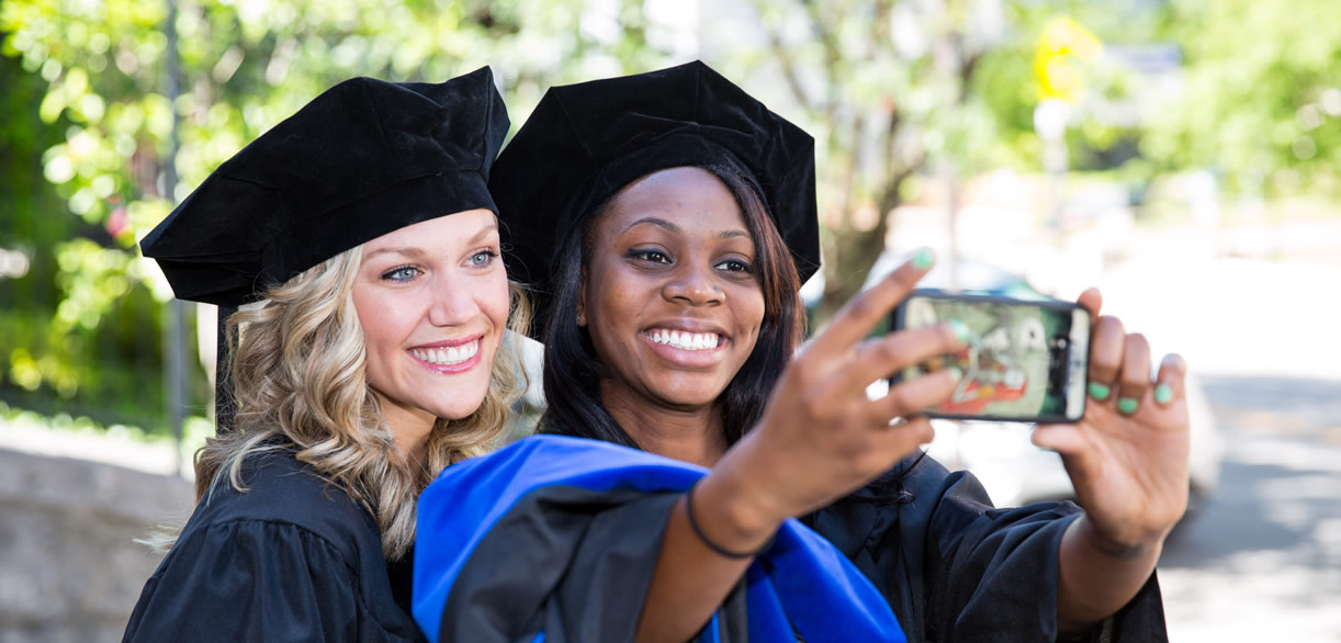 two doctor of clinical psychology graduate students in black cap and gowns take a photo at graduation