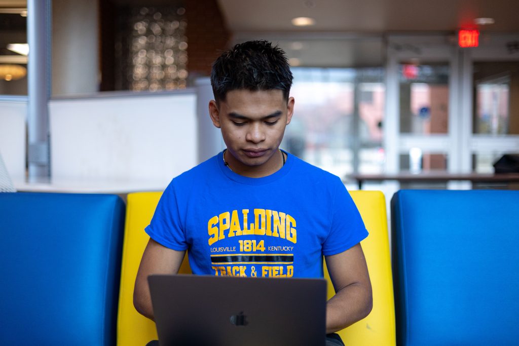 Male student in a blue and yellow Spalding t-shirt sits in a building lobby working on a laptop
