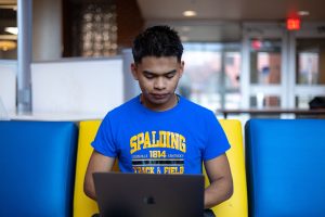 Male student in a blue and yellow Spalding t-shirt sits in a building lobby working on a laptop