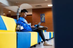 Male student in a blue and yellow Spalding t-shirt sits in a building lobby and studies on a laptop