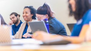 A row of four students in blue scrubs sits together with laptops in a classroom