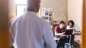 The back of a black male professor seen in a small classroom with two students studying