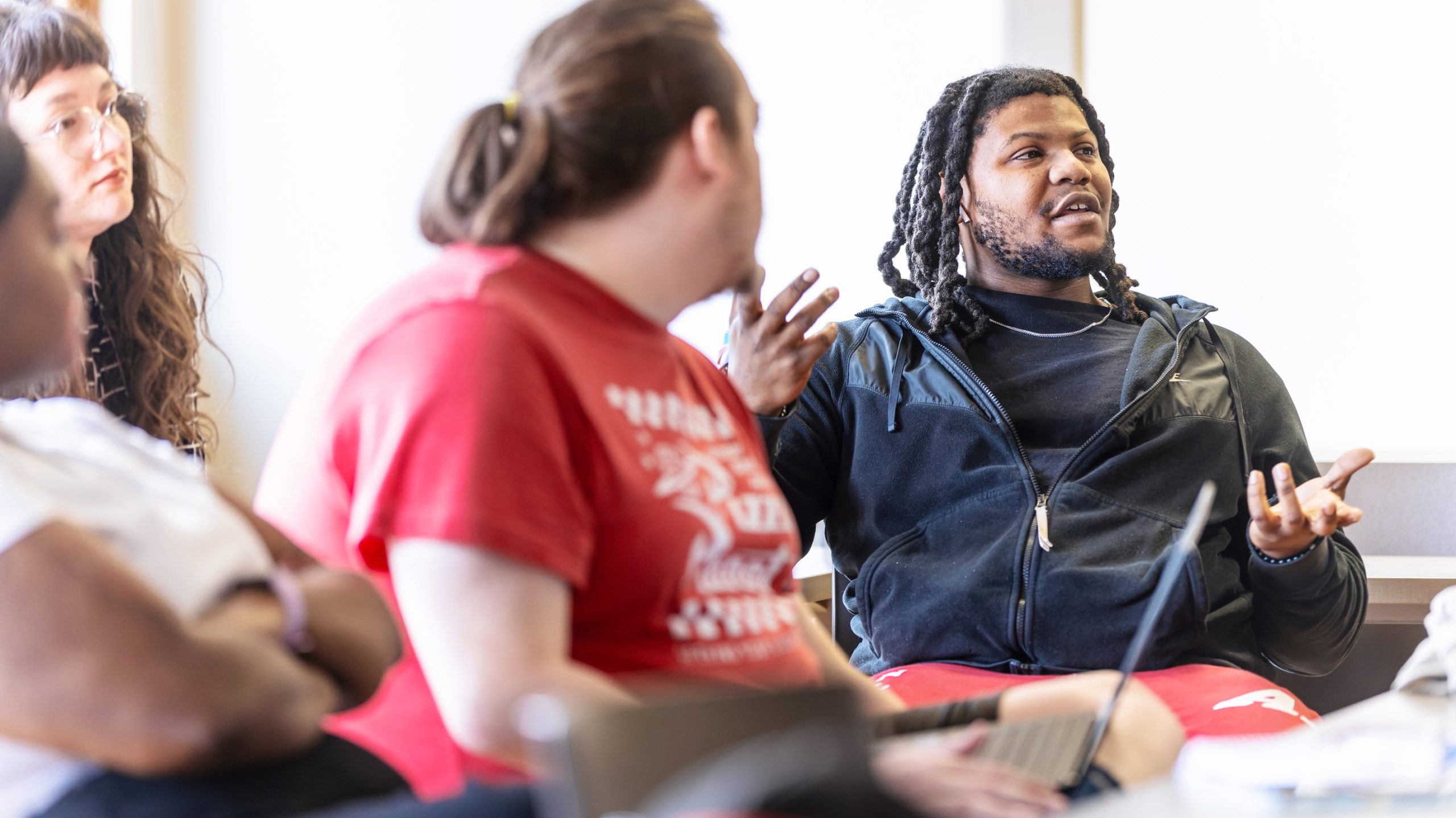 Two male students have a conversation in class while sitting with open books and computers