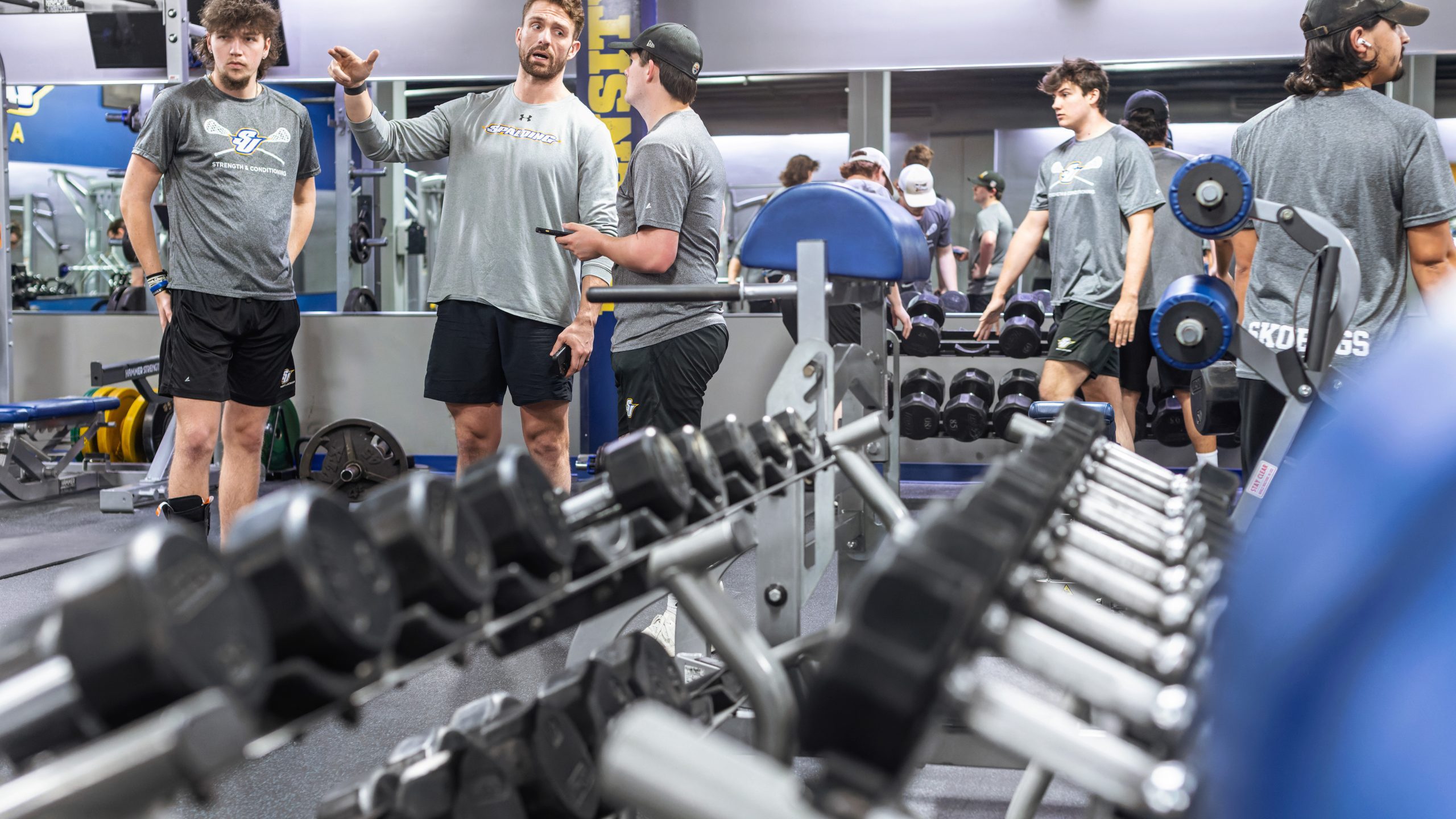 Male trainer in a long sleeve gray shirt provides feedback to two male students in the weight room