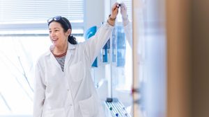 Female science professor in white lab coat stands at a board in a science classroom