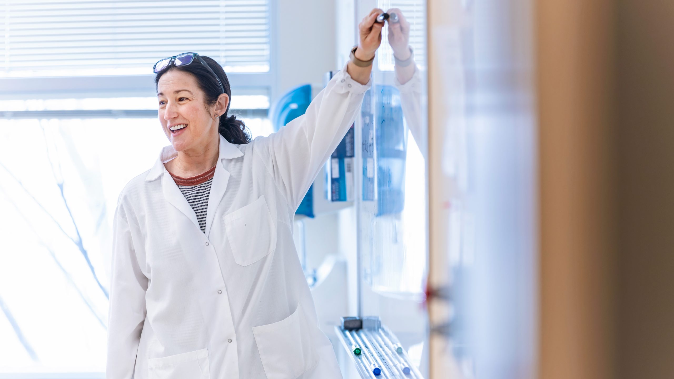 Female science professor in white lab coat stands at a board in a science classroom