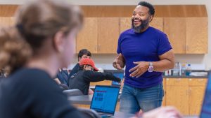 Black male professor in a blue shirt stands in a science lab talking to students