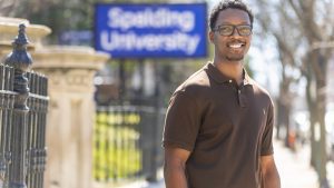Portrait of a smiling, black male student in a brown shirt and glasses