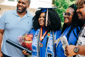 A young black woman in a blue graduation cap and gown smiles with her family at graduation