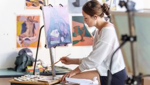 Female student in a white shirt paints at an easel in a bright studio environment