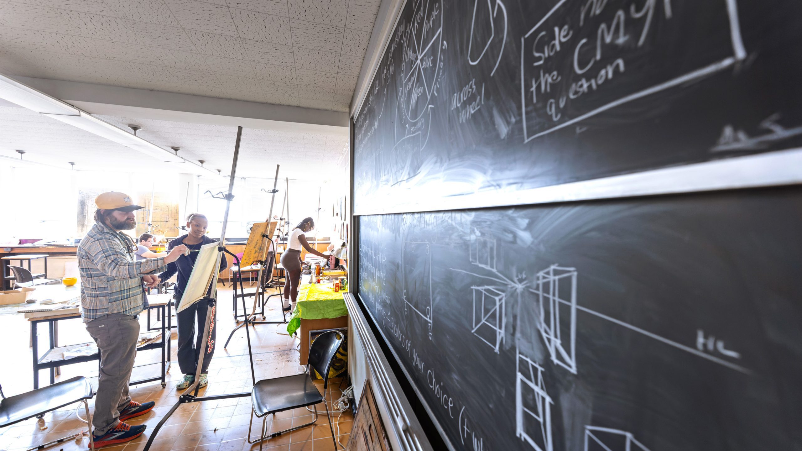 Male teacher in an open, bright classroom works with a small group of students next to a board covered in writing