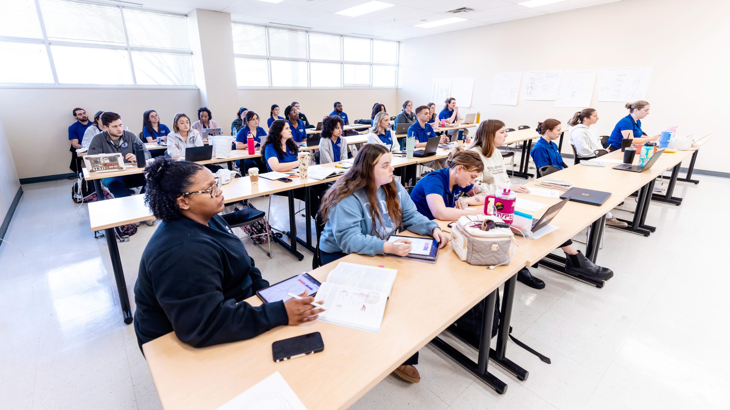 A large, bright classroom of students sit in rows of tables taking notes on tablets during a class