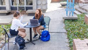 Two female students sit at a table outside with a laptop and backpacks