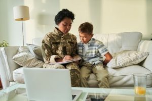 A young woman in military fatigues studies on a couch in a living room sitting next to her son
