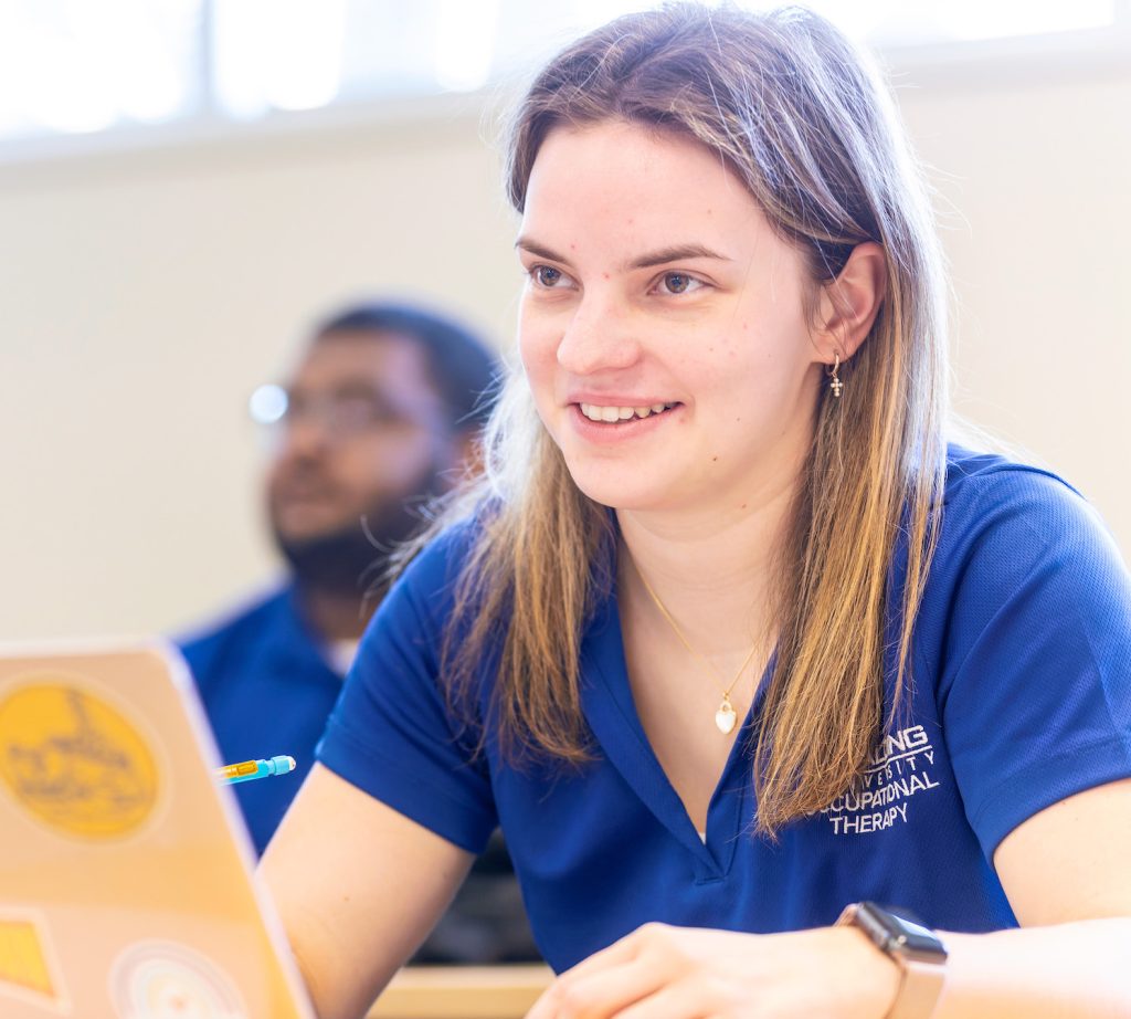 Close up of a femal student in a blue Spalding occupational therapy shirt sitting at a laptop in a classroom