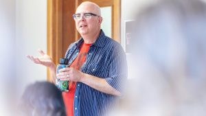 Close up of a middle age male professor in glasses, holding a water bottle while speaking in front of a classroom