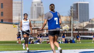 Two female Spalding student athletes hold lacrosse sticks and wear blue and white practice jerseys on an outdoor field with the city skyline in background